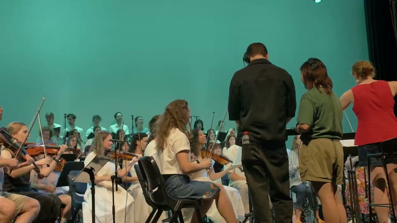 A conductor leads a youth orchestra section during a rehearsal or performance against a turquoise backdrop.