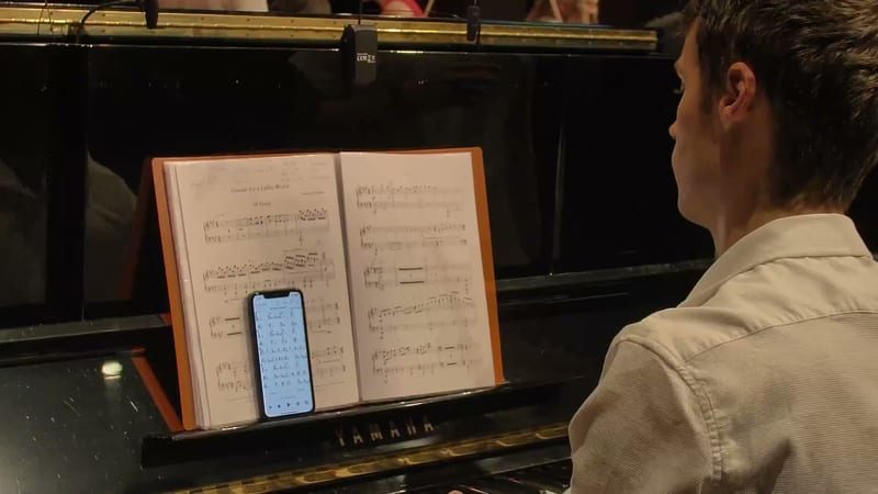 A pianist studies sheet music displayed on an orange music stand while referencing notes on a smartphone during practice.