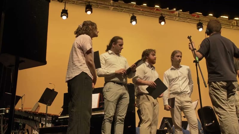 Young choir members hold sheet music while performing on stage under warm stage lighting.
