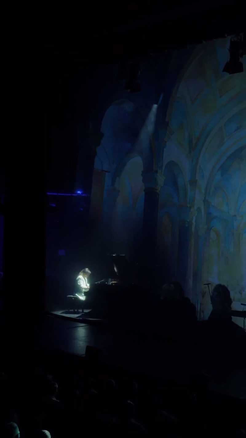 A performer plays the piano under a dramatic spotlight in front of a beautifully painted backdrop.