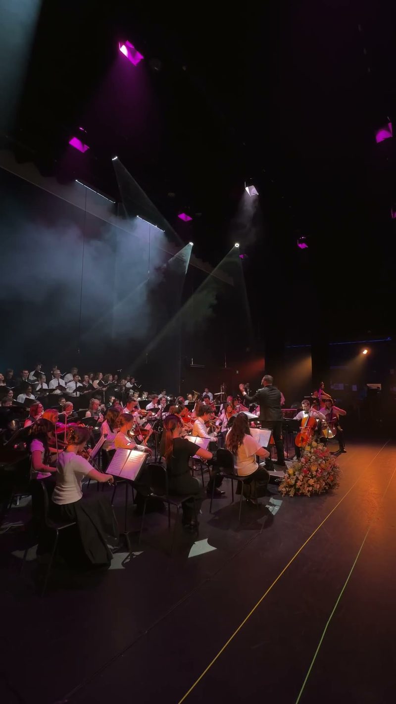 A symphony orchestra and choir perform on stage with dramatic lighting.