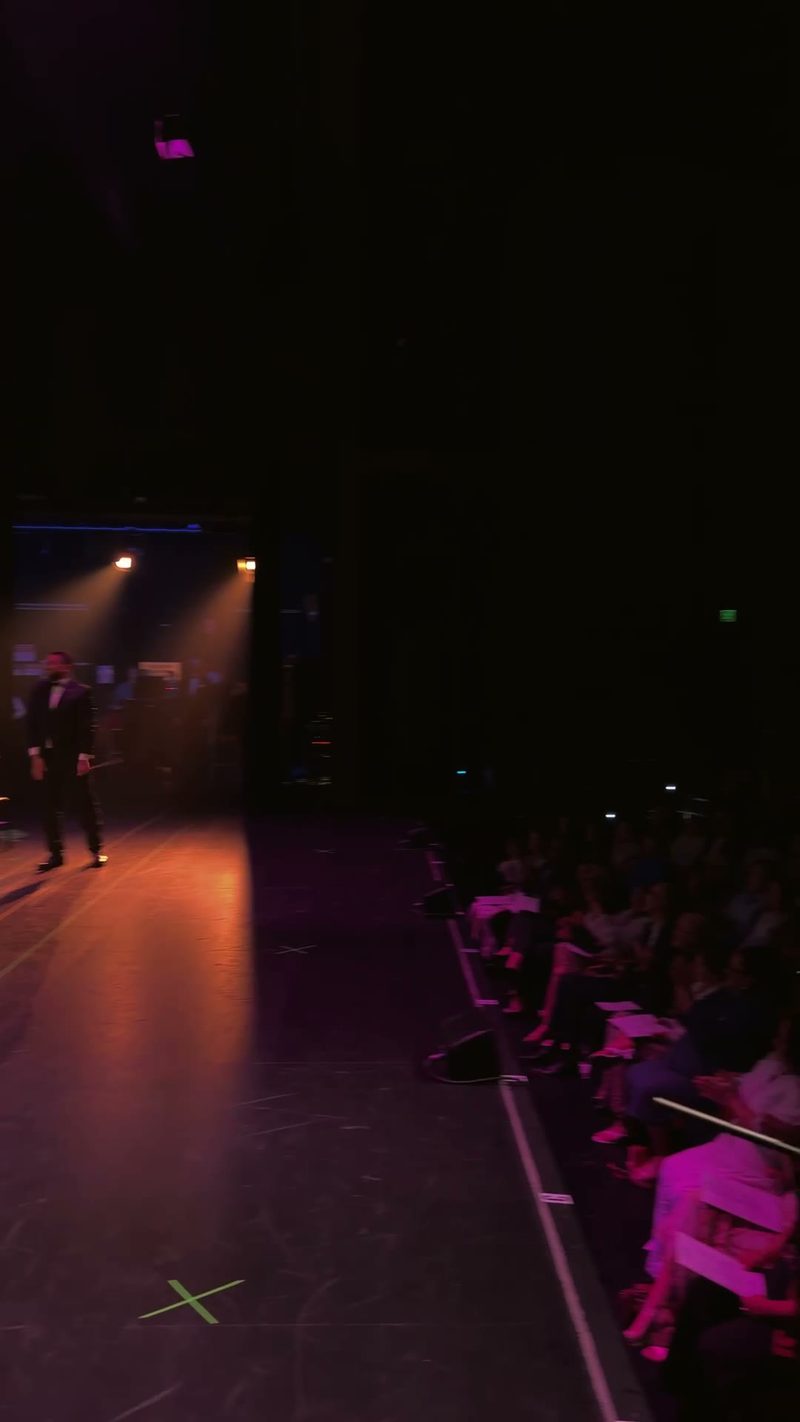 A performer wearing a tuxedo stands on stage, illuminated by spotlights, while the audience watches attentively.