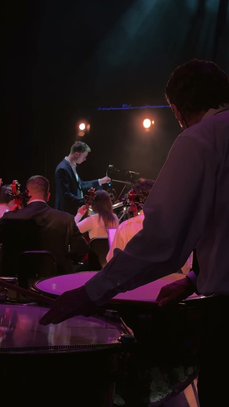 A percussionist's hand is poised above their instrument while the orchestra performs in the background.