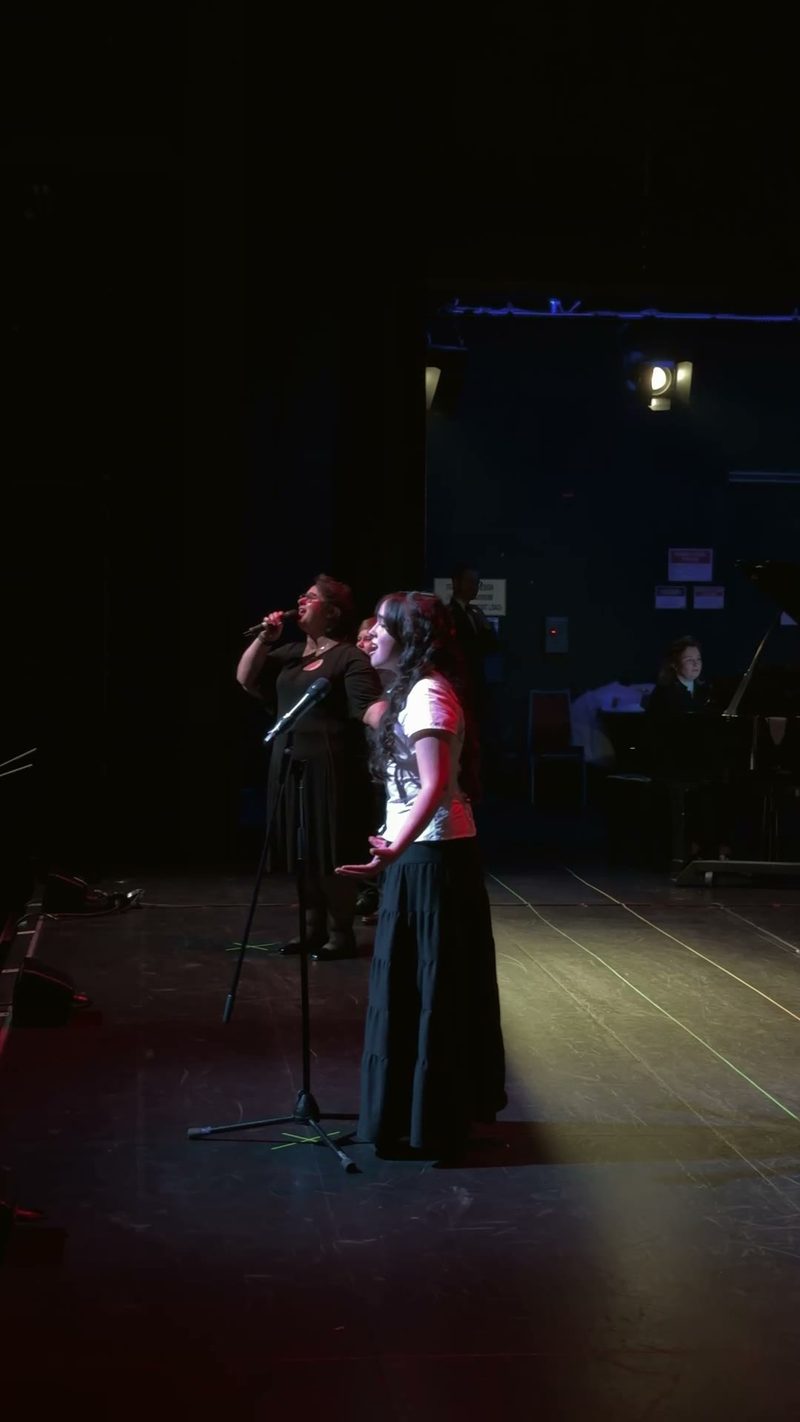 Two singers perform on stage with a pianist in the background.