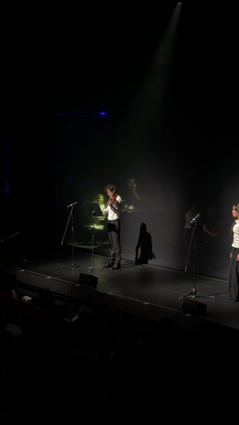 A violinist performs on stage with a pianist in the background during a live event.