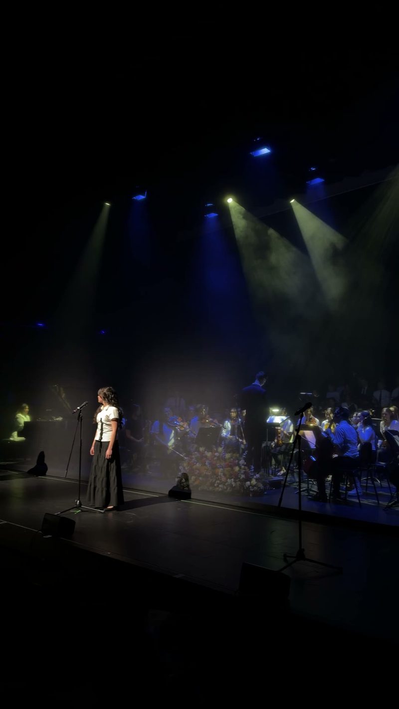 A young soloist performs on stage with a symphony orchestra in the background.