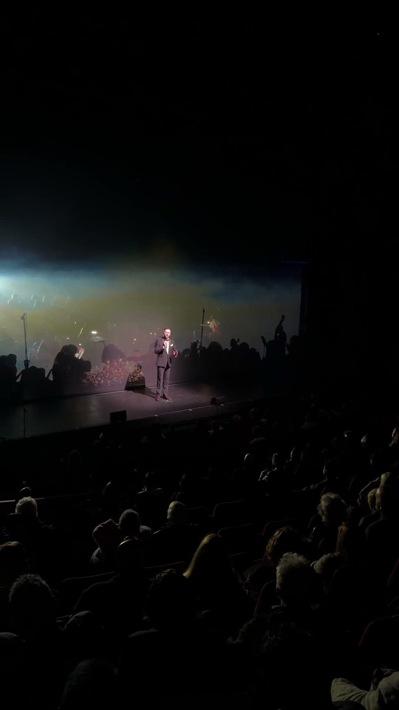 A speaker stands on stage with an orchestra visible in the background.