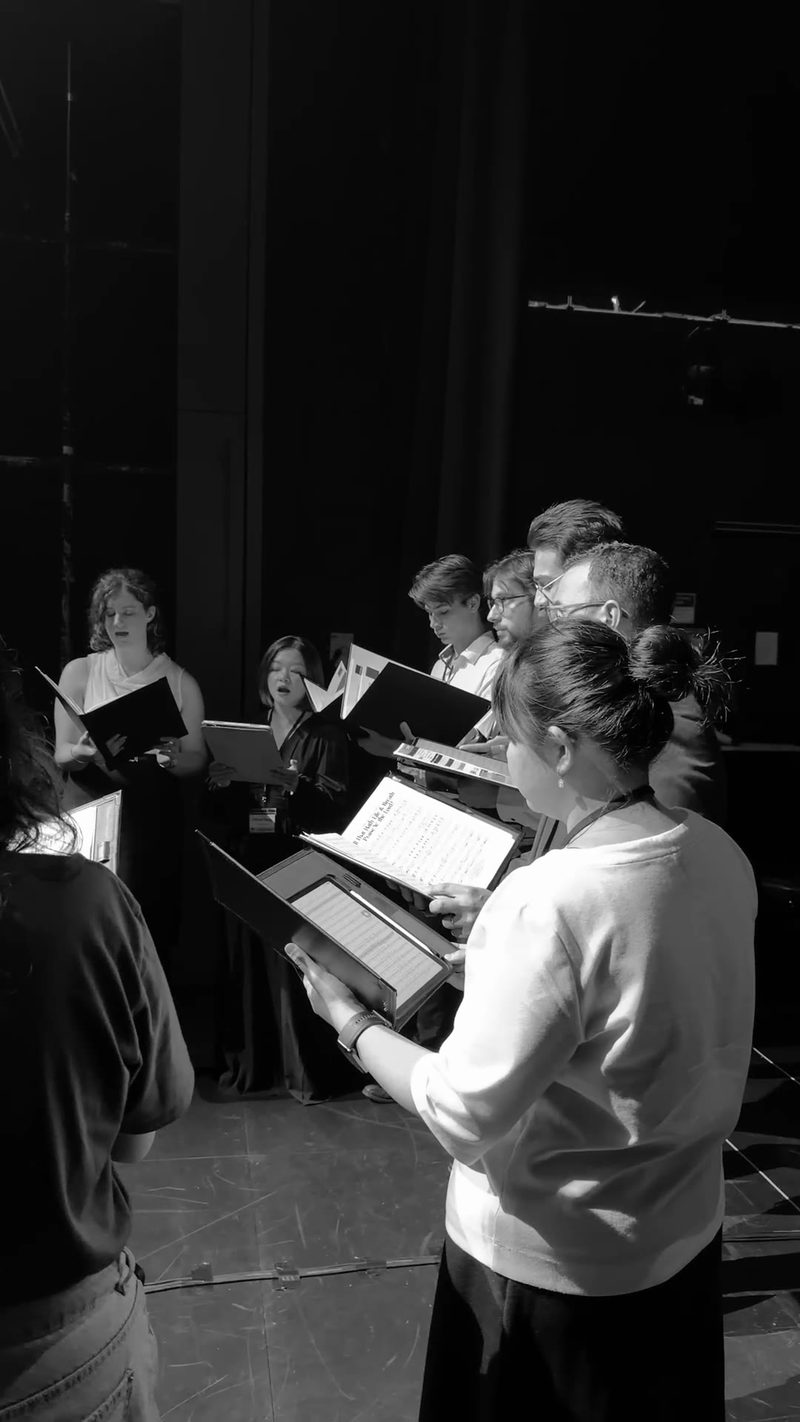 A group of singers rehearses while holding music scores in a theater setting.