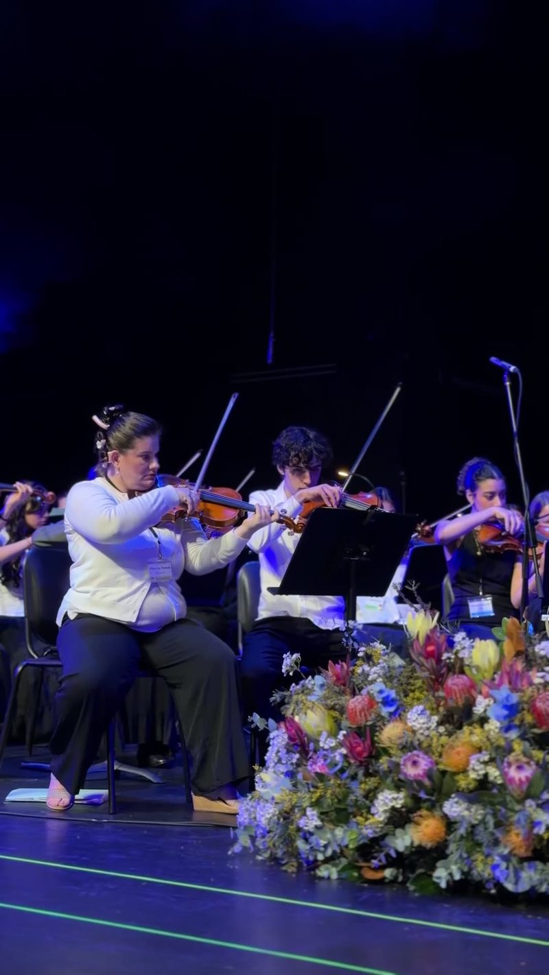 A group of musicians performing with violins on stage, surrounded by a decorative floral arrangement.