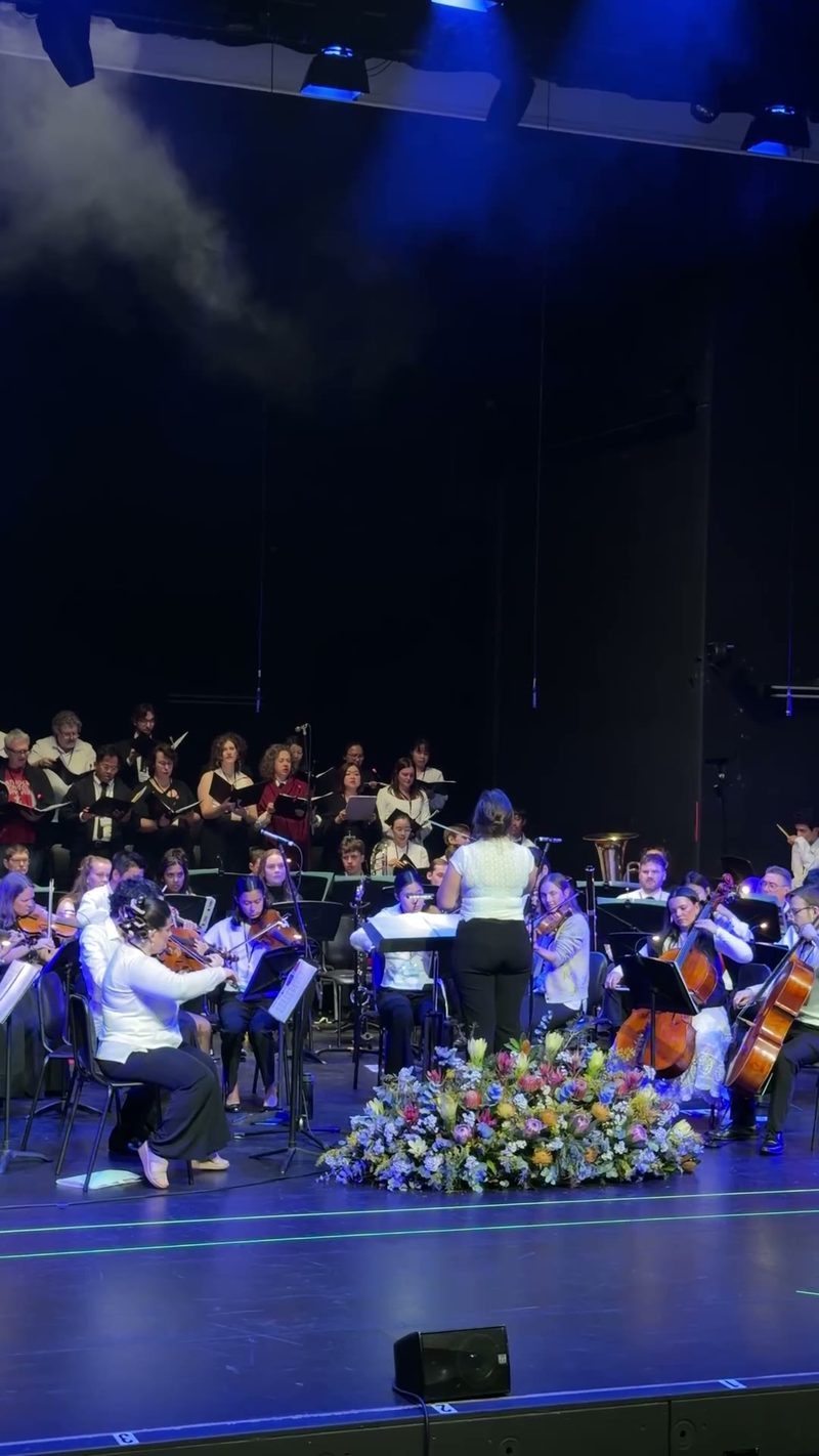 An orchestra and choir performing on stage with a floral arrangement in front.