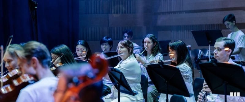 Flute and string players perform together on stage during an orchestral concert, reading from music stands under blue stage lighting.