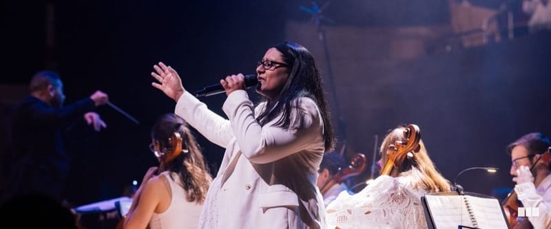 A female soloist in a white suit sings passionately into a microphone while an orchestra performs behind her and a conductor is visible in the background