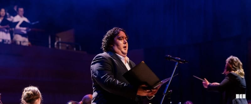 A male soloist holding a score folder sings into a microphone on a concert stage, with a conductor and choir visible in the background.
