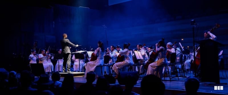 A conductor leads a full orchestra dressed in white under dramatic blue stage lighting during a concert performance.