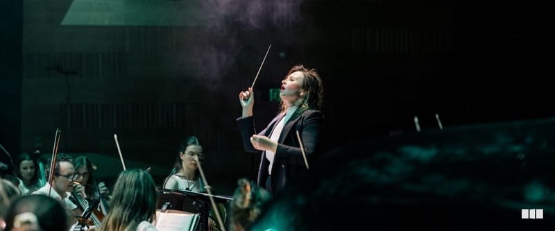 A female conductor raises her baton expressively while leading an orchestra on a dramatically lit stage.