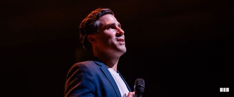 A male soloist in a navy suit looks upward while holding a microphone on a darkened stage under warm spotlight