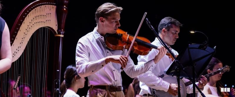 Two violinists in white shirts perform on stage with a harp visible in the background during an orchestral concert
