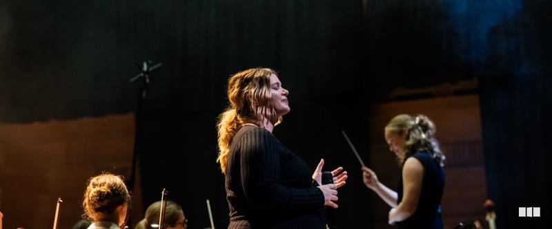 A soprano soloist performs with eyes closed and hands clasped while a conductor leads the orchestra behind her on a darkened concert stage.