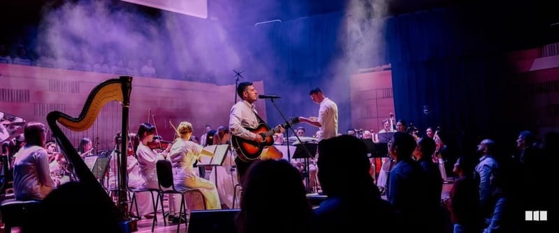 A guitarist and conductor perform at a microphone stand with a full orchestra including harp and strings in a dramatically lit concert hall.