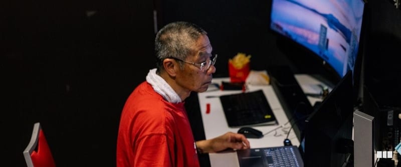 An older man wearing a red shirt and glasses works intently at a computer workstation with a large monitor in a darkened room