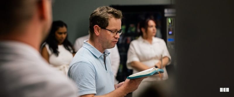A man wearing glasses reads from an open book during what appears to be a rehearsal or gathering, with other attendees visible in the background near a vending machine.