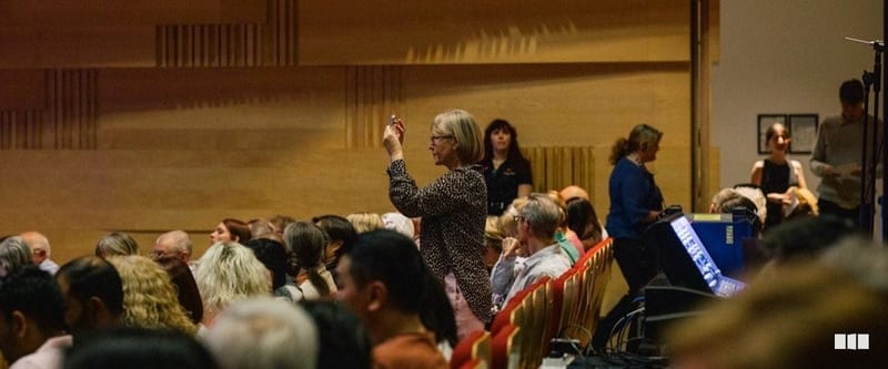 An audience member raises her hand during what appears to be a Q&A or interactive session in a warmly lit concert hall