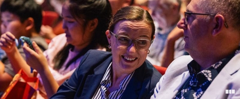 A smiling woman in glasses and a navy blazer chats with a man in a floral shirt while seated in an auditorium audience.