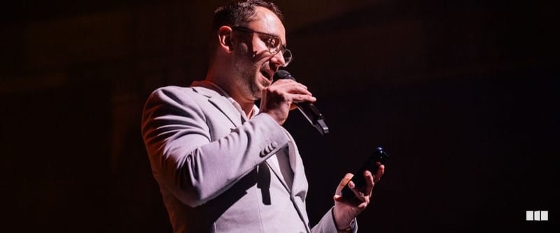A man in a light grey suit speaks energetically into a handheld microphone on a darkened stage while holding a small device in his other hand