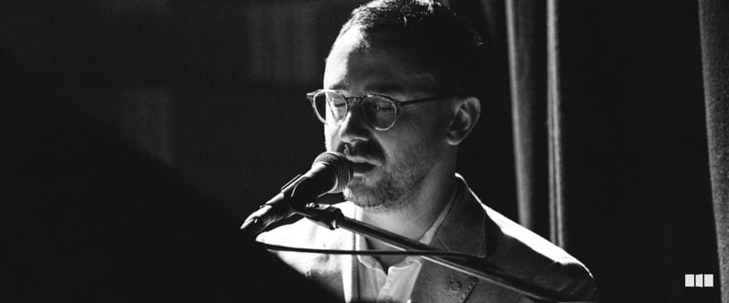 A male vocalist with glasses performs at a microphone on a dimly lit stage