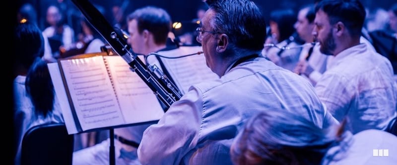 A bassoonist reads from a music stand during an orchestral performance under blue stage lighting, with fellow wind players visible behind.